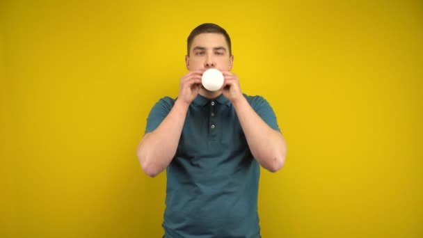 Молодий чоловік надуває білу кульку ротом на жовтому тлі. Людина в зеленому поло . — стокове відео