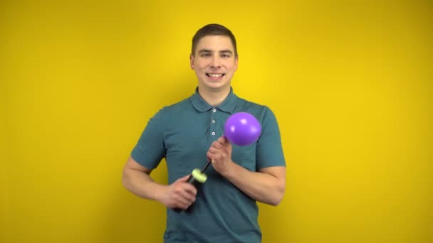 A young man inflates a purple balloon with a pump on a yellow background. Man in a green polo. — Stock Video