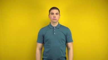 Genç bir adam, sarı arka planda parmaklarıyla neşeli bir gülümseme sergiliyor. Yeşil polo giyen adam..