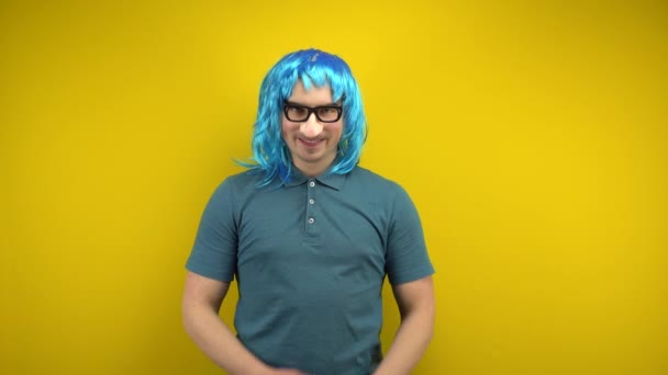Un jeune homme drôle avec une perruque bleue femelle et des lunettes avec un nez se frotte les mains et regarde autour. Tournage en studio sur fond jaune. — Video