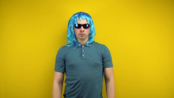 一个穿着女人蓝色假发和太阳镜的有趣的年轻人严肃地交叉着胳膊。在黄底的演播室拍摄. — 图库视频影像