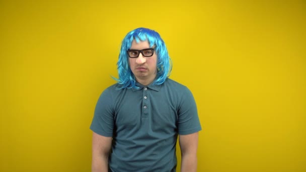 En rolig ung man i en blå peruk och glasögon med en näsa som viftar med handen. Fotografering i studion på en gul bakgrund. — Stockvideo