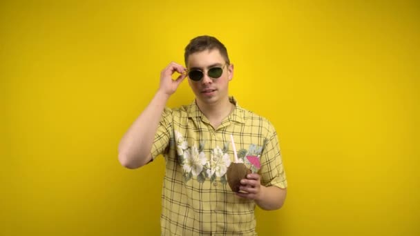 Un hombre con gafas de sol y una camisa hawaiana se quita las gafas y tiene una piña colada de coco en la mano. Tiro en el estudio sobre un fondo amarillo. — Vídeos de Stock