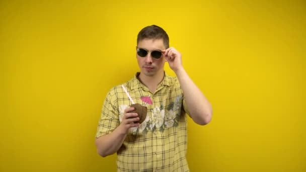 Een man met een zonnebril en een Hawaïaans shirt knipoogt en drinkt een pina colada gemaakt van kokosnoot. Opname in de studio op een gele achtergrond. — Stockvideo