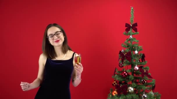 Νεαρή γυναίκα χορεύει με ένα ποτήρι σαμπάνια κοντά στο χριστουγεννιάτικο δέντρο σε κόκκινο φόντο. Ένα κορίτσι με γυαλιά και ένα βραδινό φόρεμα.. — Αρχείο Βίντεο