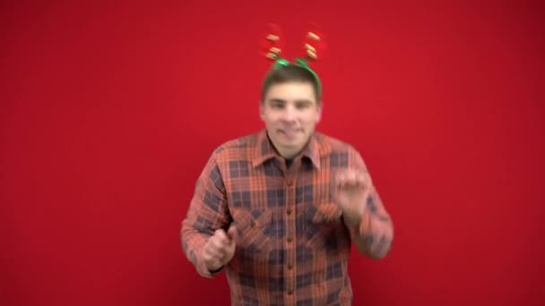 Mladý muž tančí s čelenkou v podobě vánočních parohů. Studio natáčení na červeném pozadí. — Stock video