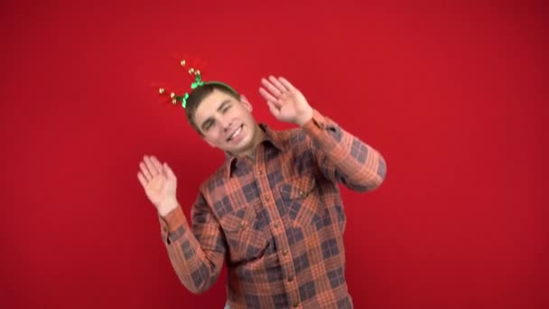 Mladý muž tančí s čelenkou v podobě vánočních parohů. Studio natáčení na červeném pozadí. — Stock video
