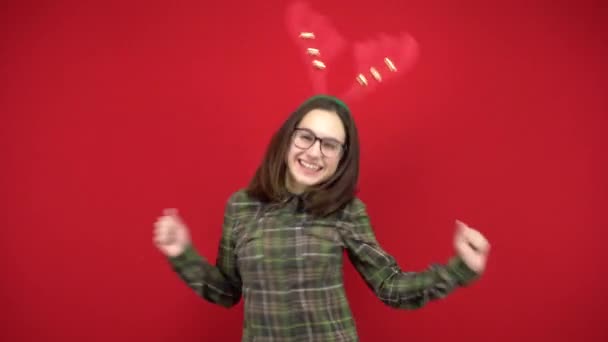 En ung kvinna dansar med ett pannband i form av julhorn. Studio fotografering på en röd bakgrund. — Stockvideo