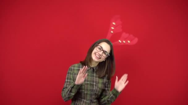 En ung kvinna dansar med ett pannband i form av julhorn. Studio fotografering på en röd bakgrund. — Stockvideo