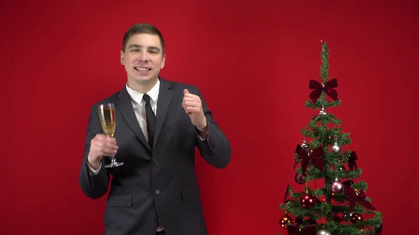 Genç adam kırmızı arka planda Noel ağacının yanında bir kadeh şampanyayla dans ediyor. Resmi takım elbiseli bir adam.. — Stok fotoğraf