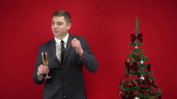Genç adam kırmızı arka planda Noel ağacının yanında bir kadeh şampanyayla dans ediyor. Resmi takım elbiseli bir adam.. — Stok fotoğraf