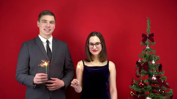 Ein Liebespaar hält Wunderkerzen am Weihnachtsbaum in den Händen. Junger Mann und Frau im Atelier auf rotem Hintergrund. — Stockfoto