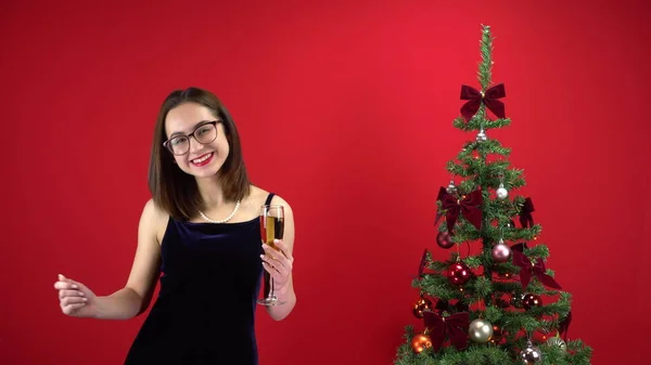 Egy fiatal nő táncol egy pohár pezsgővel a karácsonyfa mellett, piros háttérrel. Egy szemüveges lány és egy estélyi ruha.. — Stock Fotó