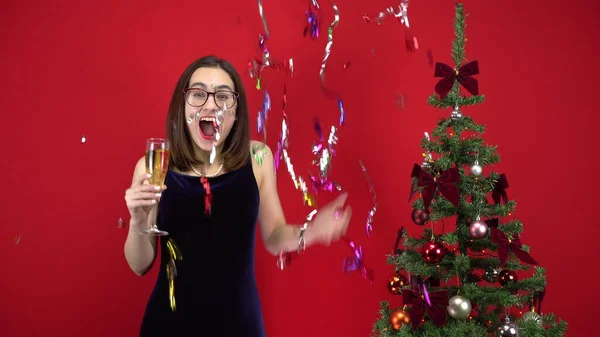 Mujer joven cuenta cinco segundos para la fiesta con una copa de champán cerca del árbol de Navidad sobre un fondo rojo. Una chica con gafas y un vestido de noche. —  Fotos de Stock