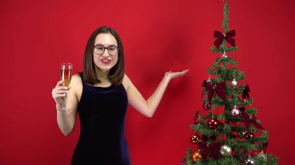 La jeune femme compte cinq secondes avant la fête avec une coupe de champagne près du sapin de Noël sur un fond rouge. Une fille avec des lunettes et une robe de soirée. — Photo