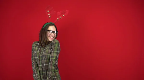 Une jeune femme se tient avec un bandeau en forme de cornes de Noël et est timide. Photographie studio sur fond rouge. — Photo