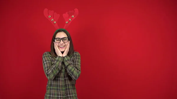 Une jeune femme se tient avec un bandeau en forme de cornes de Noël et est timide. Photographie studio sur fond rouge. — Photo