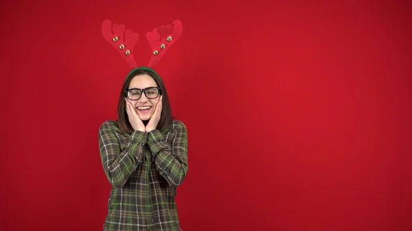 Una mujer joven se para con una diadema en forma de cuernos de Navidad y es tímida. Fotografía de estudio sobre fondo rojo. —  Fotos de Stock