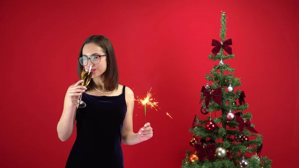 O tânără stă cu un pahar de șampanie și un strălucitor lângă pomul de Crăciun pe un fundal roșu. O fată cu ochelari și o rochie de seară . — Fotografie, imagine de stoc