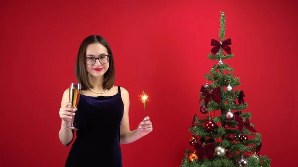 Una joven está de pie con una copa de champán y una espumosa cerca del árbol de Navidad sobre un fondo rojo. Una chica con gafas y un vestido de noche. —  Fotos de Stock