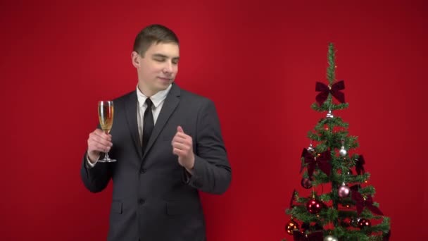 Ung man dansar med ett glas champagne nära julgranen på en röd bakgrund. En man i en formell kostym. — Stockvideo