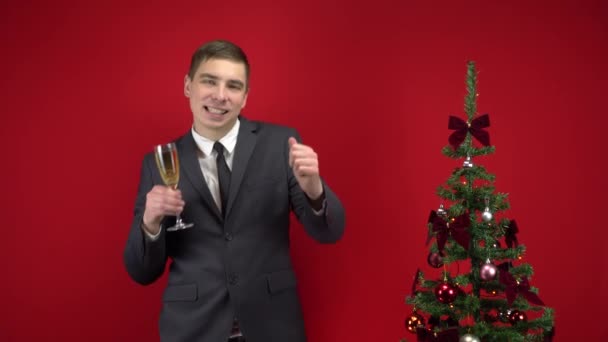 Jeune homme dansant avec une coupe de champagne près du sapin de Noël sur fond rouge. Un homme en costume officiel. — Video