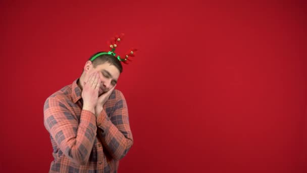 Mladý muž stojí s čelenkou v podobě vánočních rohů a stydí se. Studio fotografie na červeném pozadí. — Stock video