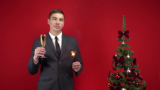 젊은 남자가 샴페인 한 잔과 작은 스파클러 한 잔을 들고 빨간 배경의 크리스마스 트리 근처에 서 있다. 정장을 입은 남자. — 비디오