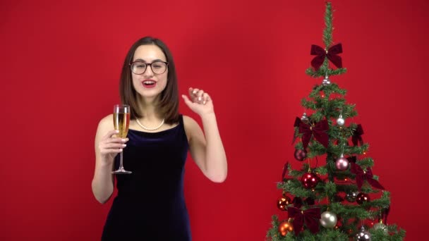 A jovem conta cinco segundos para o feriado com uma taça de champanhe perto da árvore de Natal em um fundo vermelho. Uma menina com óculos e um vestido de noite. — Vídeo de Stock