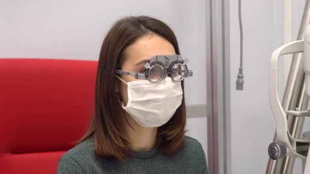 Eine junge Frau sitzt mit einer Spezialbrille zur Auswahl der nötigen Linsen beim Augenarzt. Das Mädchen trägt eine Schutzmaske vor dem Virus. — Stockvideo
