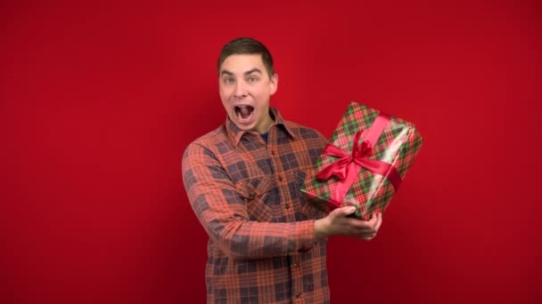 Egy fiatalember megrázza a karácsonyi ajándékot a kezében, és örül. Fényképezés a stúdióban a piros háttér. — Stock videók