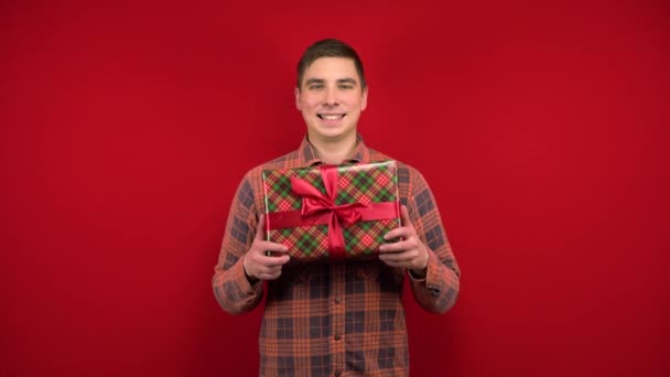 Ein junger Mann hält ein Weihnachtsgeschenk in der Hand und freut sich. Dreharbeiten im Studio auf rotem Hintergrund. — Stockvideo