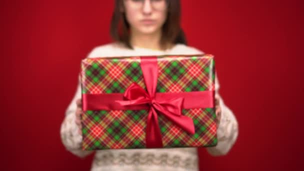 Μια νεαρή γυναίκα κρατάει ένα χριστουγεννιάτικο δώρο στην κάμερα. Πυροβολισμοί στο στούντιο σε κόκκινο φόντο. — Αρχείο Βίντεο