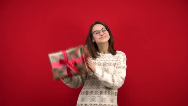 Una joven con gafas se sacude un regalo de Navidad en sus manos y se regocija. Tiro en el estudio sobre un fondo rojo. — Vídeos de Stock