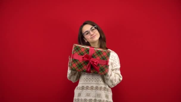 Een jonge vrouw met een bril schudt een kerstcadeau in haar handen en verheugt zich. Opname in de studio op een rode achtergrond. — Stockvideo