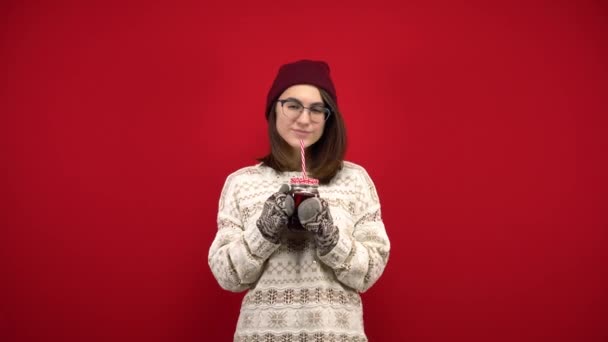 A young woman in glasses, a hat and mittens drinks mulled wine. Shooting in the studio on a red background. — Stock Video