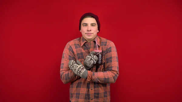 모자를 쓰고 벙어리장갑을 끼고 있는 젊은 남자가 계속 얼어 있고 다진 포도주를 마십니다. 붉은 배경으로 스튜디오에서 촬영. — 스톡 사진