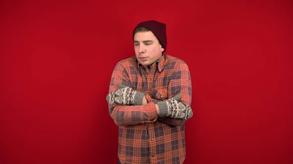 Un joven con una camisa roja, sombrero y manoplas es frío y se abraza para mantenerse caliente. Tiro en el estudio sobre un fondo rojo. —  Fotos de Stock