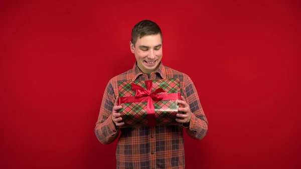 Genç bir adam elinde bir Noel hediyesi sallıyor ve seviniyor. Stüdyoda kırmızı arka planda çekim yapmak. — Stok fotoğraf