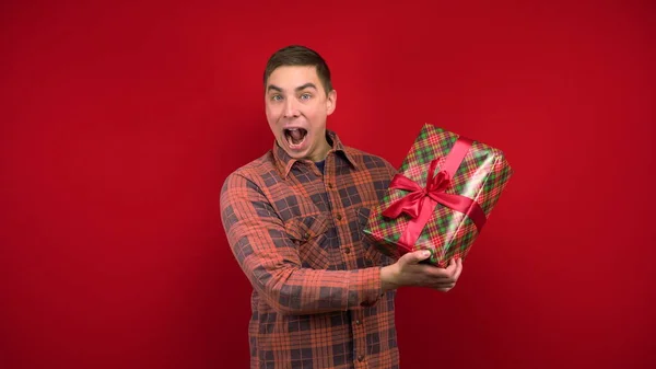 Genç bir adam elinde bir Noel hediyesi sallıyor ve seviniyor. Stüdyoda kırmızı arka planda çekim yapmak. — Stok fotoğraf