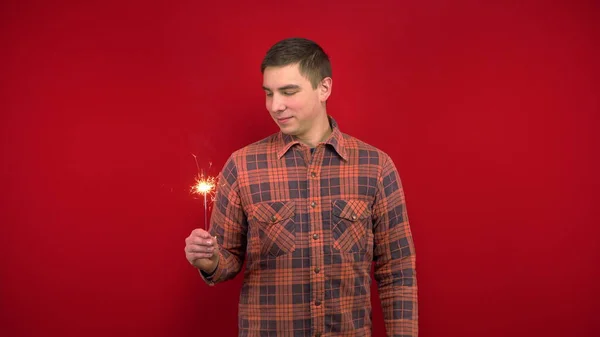 Ein junger Mann im roten Hemd hält eine Wunderkerze in der Hand. Dreharbeiten im Studio auf rotem Hintergrund. — Stockfoto