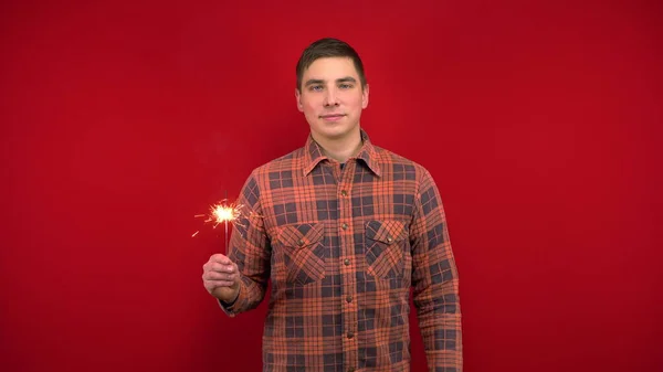 Ein junger Mann im roten Hemd hält eine Wunderkerze in der Hand. Dreharbeiten im Studio auf rotem Hintergrund. — Stockfoto