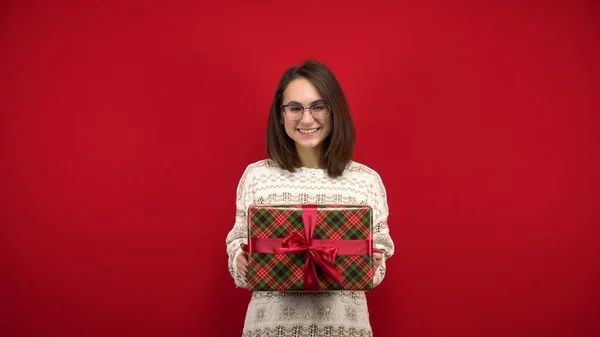 O tânără cu ochelari scutură un cadou de Crăciun în mâini și se bucură. Impuscaturi in studio pe un fundal rosu . — Fotografie, imagine de stoc