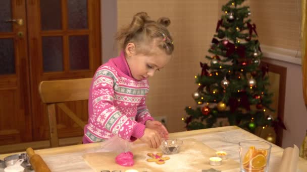 Una ragazza decora un biscotto a forma umana di pan di zenzero con glassa rosa. Il bambino si sta preparando per il Natale sullo sfondo dell'albero di Natale. — Video Stock