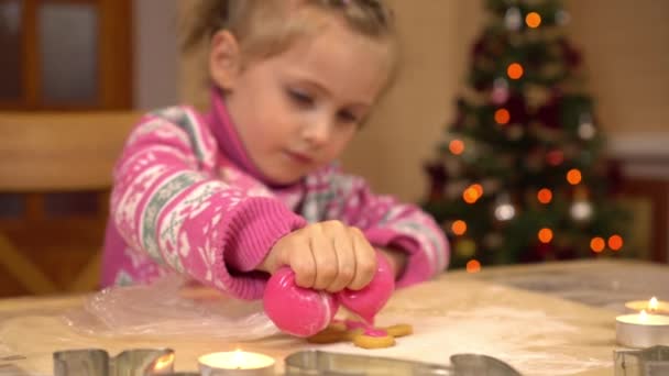 En flicka dekorerar en pepparkaka människoformad kaka med rosa glasyr. Barnet förbereder sig för jul på bakgrunden av julgranen. — Stockvideo