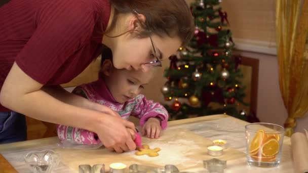 Uma jovem mãe ajuda sua filha a decorar biscoitos de gengibre na forma de um humano com cobertura. A criança está se preparando para o Natal no fundo da árvore de Natal. — Vídeo de Stock