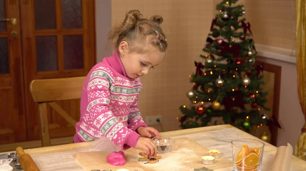 한 소녀는 진저브레드 인간 모양의 쿠키를 분홍색으로 칠하고 있다. 아이는 크리스마스 트리를 배경으로 준비하고 있습니다.. — 스톡 사진