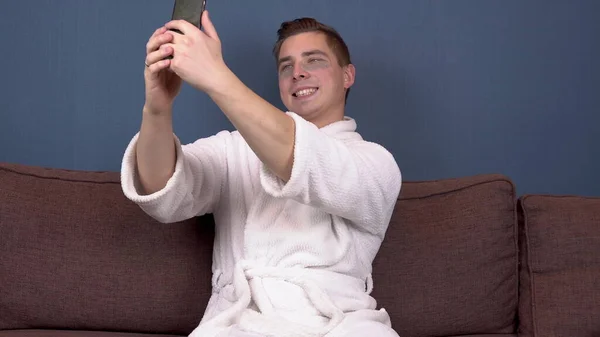 Ein junger Mann mit Pflaster im Gesicht macht ein Selfie. Blaue Hydrogelpflaster zur Gesichtsverjüngung. Ein Mann sitzt im weißen Mantel auf der Couch. — Stockfoto