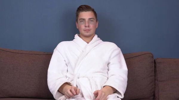 Un joven con manchas en la cara se relaja. Tratamientos de spa. Parches de hidrogel azul para el rejuvenecimiento facial. Un hombre sentado en un abrigo blanco en el sofá. — Foto de Stock