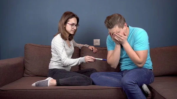 一位年轻妇女向一位年轻丈夫显示出怀孕测试呈阳性。 这个人很不高兴,抓住了他的头. 夫妇俩坐在沙发上彼此相对 — 图库照片